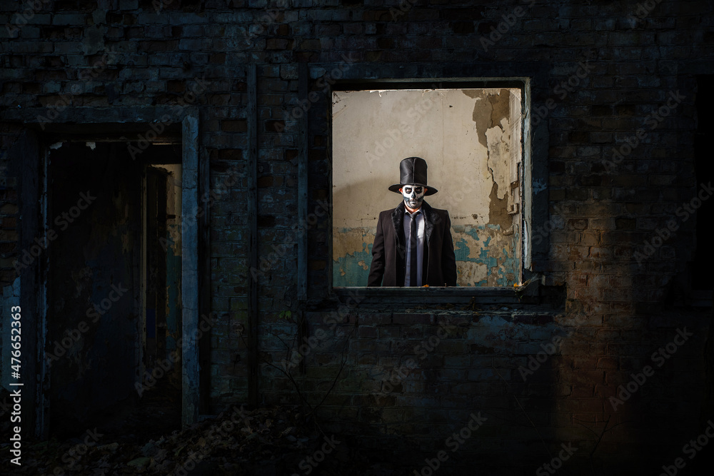 A man with skull make-up in the form of death in a tailcoat and a top hat near the ruins of an old building. The day of the Dead. Halloween. A man with skull make-up in the form of death in a tailcoat and a top hat near the ruins of an old building. The day of the Dead. Halloween.