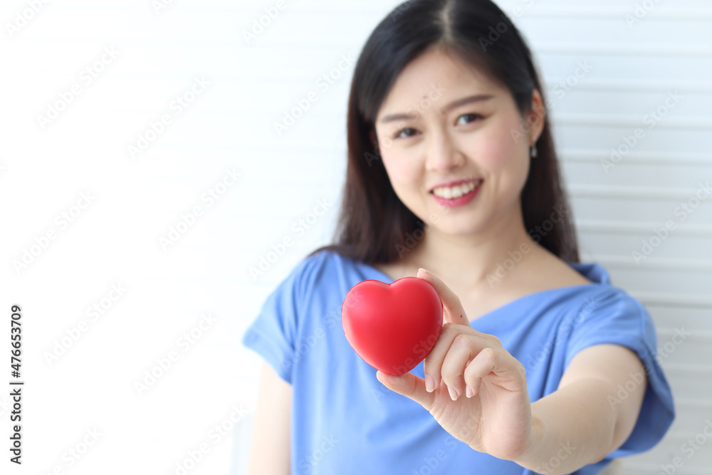 Close up of red heart on woman hand with blurred background of happy smiling female wearing blue shirt on white wall. Health, medicine and charity concept Close up of red heart on woman hand with blurred background of happy smiling female wearing blue shirt on white wall. Health, medicine and charity concept
