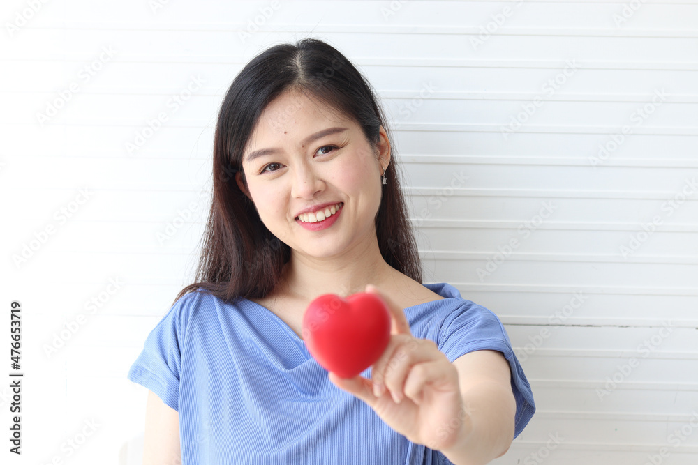 Happy smiling beautiful woman wearing blue shirt holding red heart on white wall. Health, medicine and charity concept Happy smiling beautiful woman wearing blue shirt holding red heart on white wall. Health, medicine and charity concept