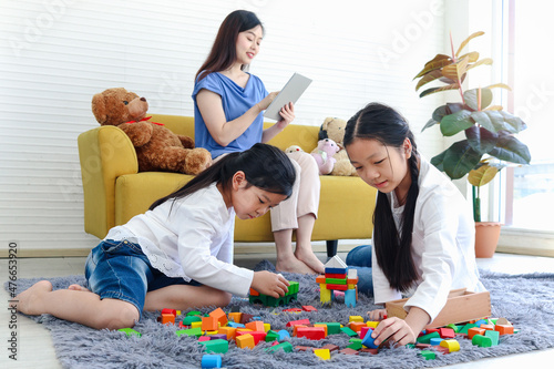 Two daughter girls playing with wooden block game (Jenga) together while mother sitting on sofa and surfing the internet, mom online working by using tablet, sister kid spending time together.