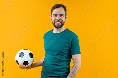 Portrait of young handsome bearded soccer supporter man posing with a ball in hand, isolated over yellow background