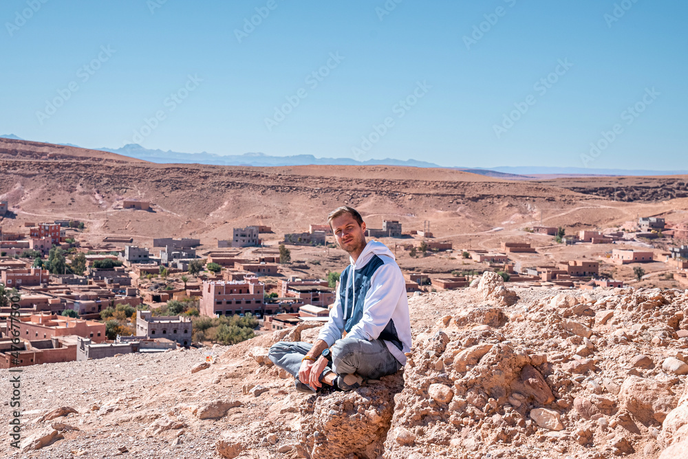 Young man sitting on rock against scenic view of rural settlements on deserted land and cloudy sky