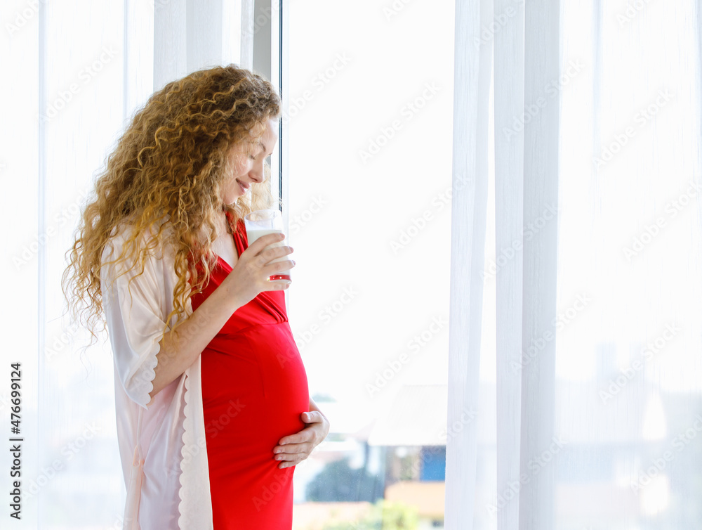 Caucasian curly long hairstyle pregnant female mother in red maternity dress pajama with rope standing smiling in front of curtain holding hand on big tummy belly with love drinking milk in glass