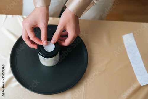 Close-up high-angle view of unrecognizable female artisan putting sticker on glass vessel with handmade candles. Process of making handmade natural candle at workshop.