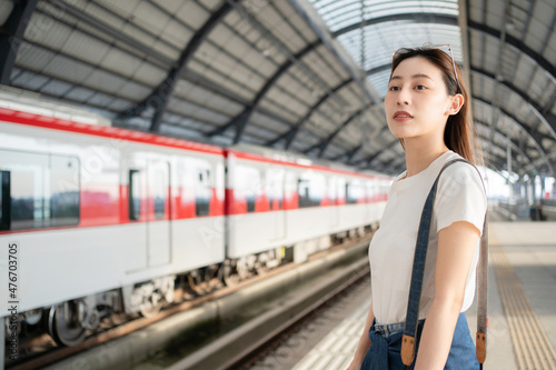 Selective focus of a happy Asian female traveler carrying a camera standing waiting for an arrival of skytrain on a platform with a blurred background of train in opposite side at a skytrain station.