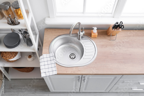 Wooden counter with silver sink, detergent and napkin near window in kitchen