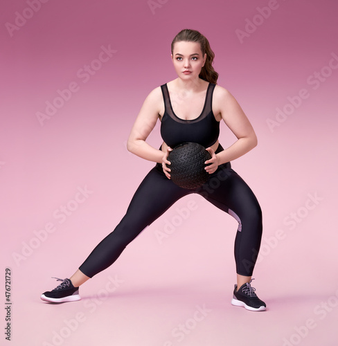 Sporty woman doing exercise with medicine ball. Photo of model with curvy figure in fashionable sportswear on pink background. Sports motivation and healthy lifestyle