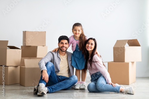 Cheerful eastern family of three people moving to new house, sitting among cardboard boxes in empty living room