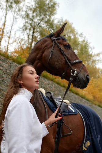 Female horseman going with her brown horse outdoor. Concept of animal care. Rural rest and leisure. Idea of green tourism. Young european woman wearing helmet and uniform © Georgii