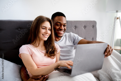 Happy young multinational couple relaxing on bed, using laptop, watching movies together