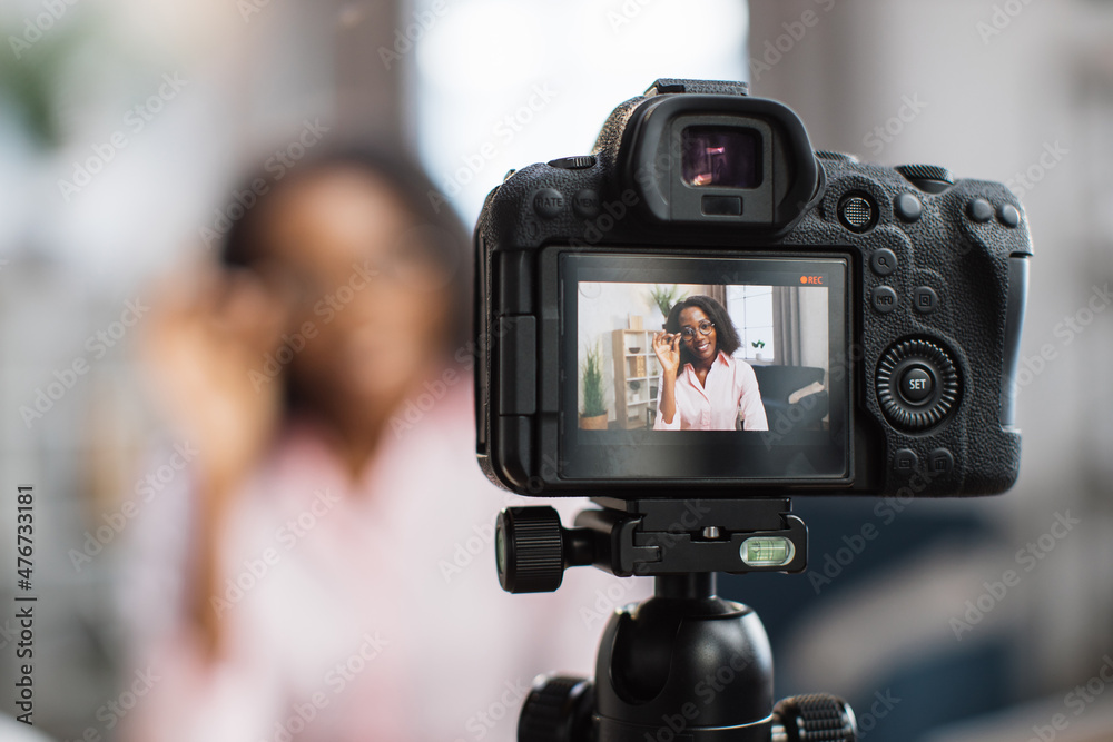 Smiling african woman talking and gesturing in front of modern video camera while sitting on blur background. Live streaming concept.