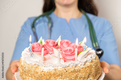 A woman doctor in a blue uniform holds a birthday cake with candles and smiles.