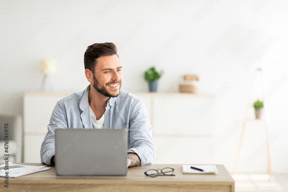 Happy freelance man working on laptop in home office, sitting on workplace and looking aside at free space Happy freelance man working on laptop in home office, sitting on workplace and looking aside at free space