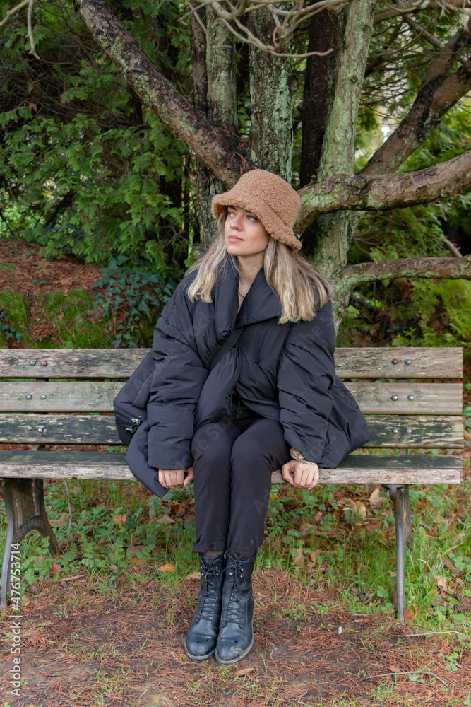 A girl in black is sitting on a bench under tree. A lonely, single, cute, beautiful model. She wears black coat and black boots. She has hat. Beauty and winter fashion concept. A girl in black is sitting on a bench under tree. A lonely, single, cute, beautiful model. She wears black coat and black boots. She has hat. Beauty and winter fashion concept.