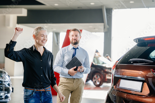 The successful visit to the dealership. Happy middle age blond woman dancing while chooses and buying a new car for herself.