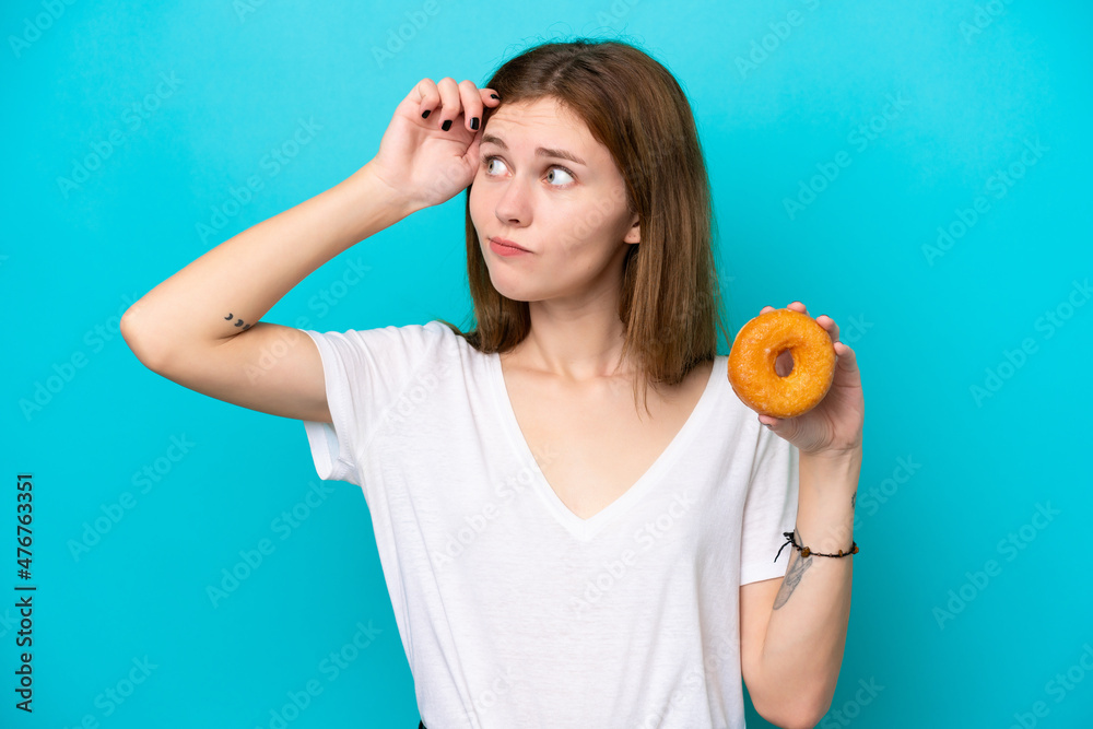 Young English woman holding a donut over isolated blue background having doubts and with confuse face expression