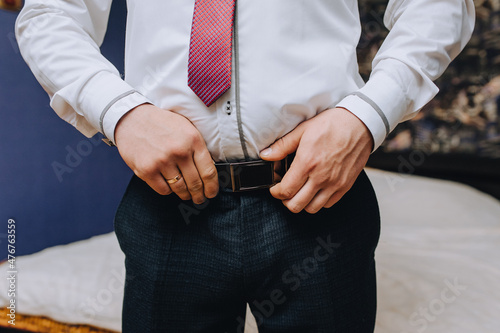 A man, a groom in a white shirt and a red tie puts a leather belt on black trousers, going to a wedding, wedding in the morning.