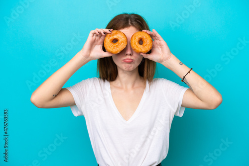 Young English woman isolated on blue background holding donuts in eyes with sad expression