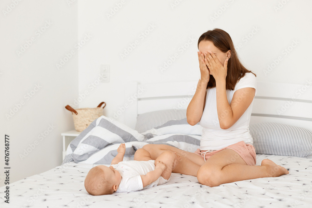 Ταπετσαρία Indoor shot of happy positive woman wearing white casual t shirt posing in bedroom with infant baby, mother covering eyes with palms, playing with child peekaboo