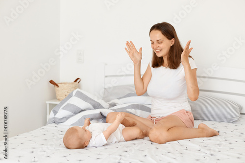 Foto Horizontal shot of happy playful mother wearing white casual t shirt sitting on bed in bedroom with toddler baby, playing hide-and-seek with her little child