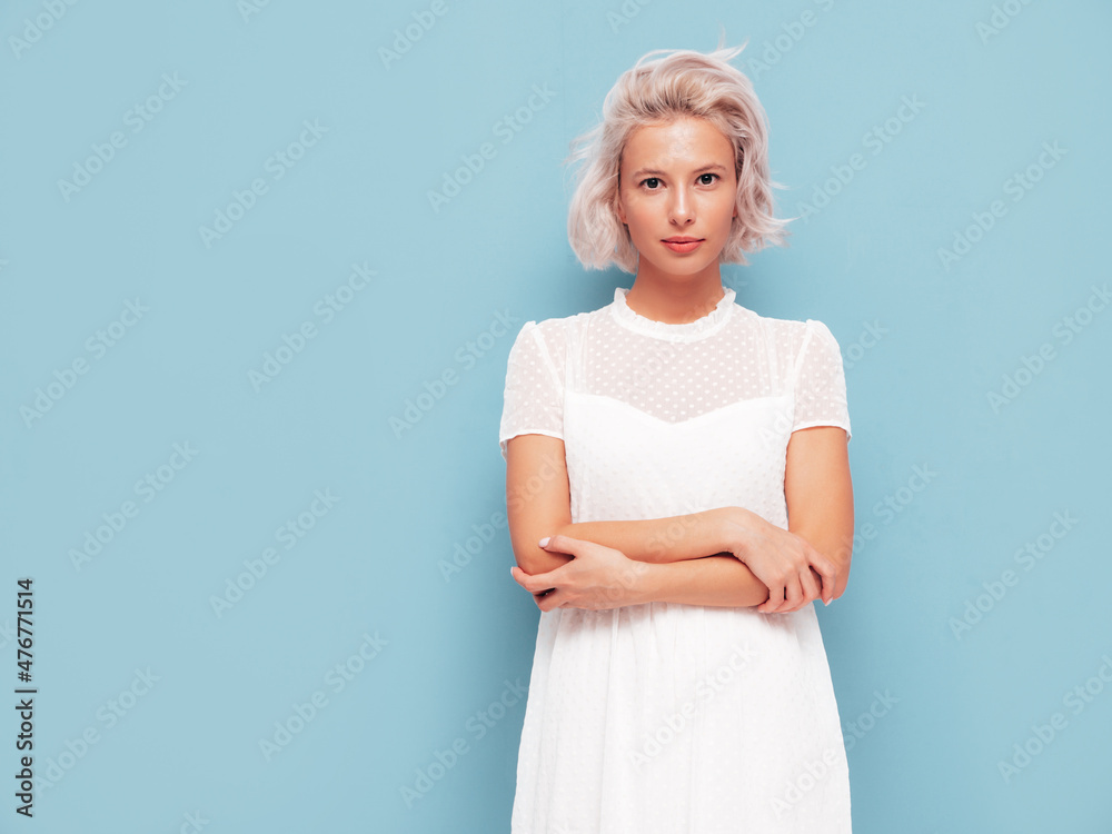 Portrait of young beautiful smiling female in trendy summer white dress. Sexy carefree blond woman posing near blue wall in studio. Positive model having fun indoors. Cheerful and happy Portrait of young beautiful smiling female in trendy summer white dress. Sexy carefree blond woman posing near blue wall in studio. Positive model having fun indoors. Cheerful and happy