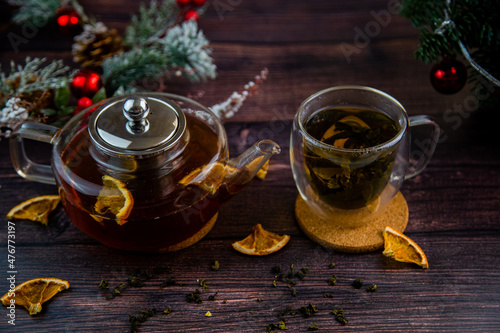 A transparent teapot and a cup of tea stand against a background of dark wood, slices of dried orange and Christmas decorations. High quality photo