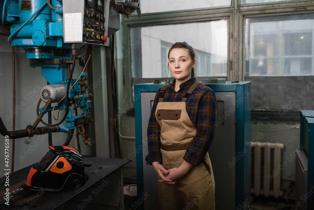 Welder in overalls standing near welding machine and protective mask in factory Welder in overalls standing near welding machine and protective mask in factory