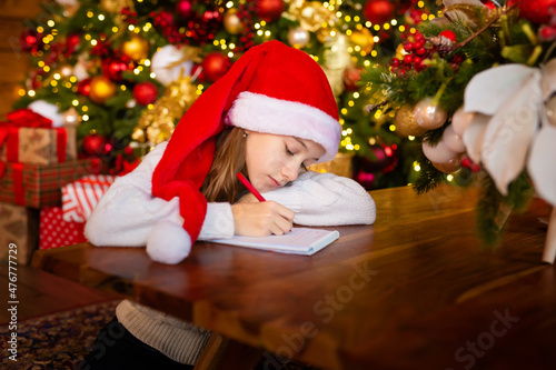 Merry Christmas and Happy Holidays. little child girl in santa claus cap writes letter Santa Claus and dreams of a gift background New Year tree indoors. Greeting card, Christmas mood concept.