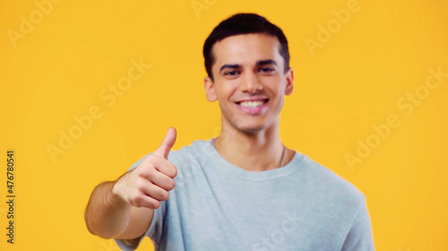 smiling and blurred man looking at camera while showing thumb up isolated on yellow.