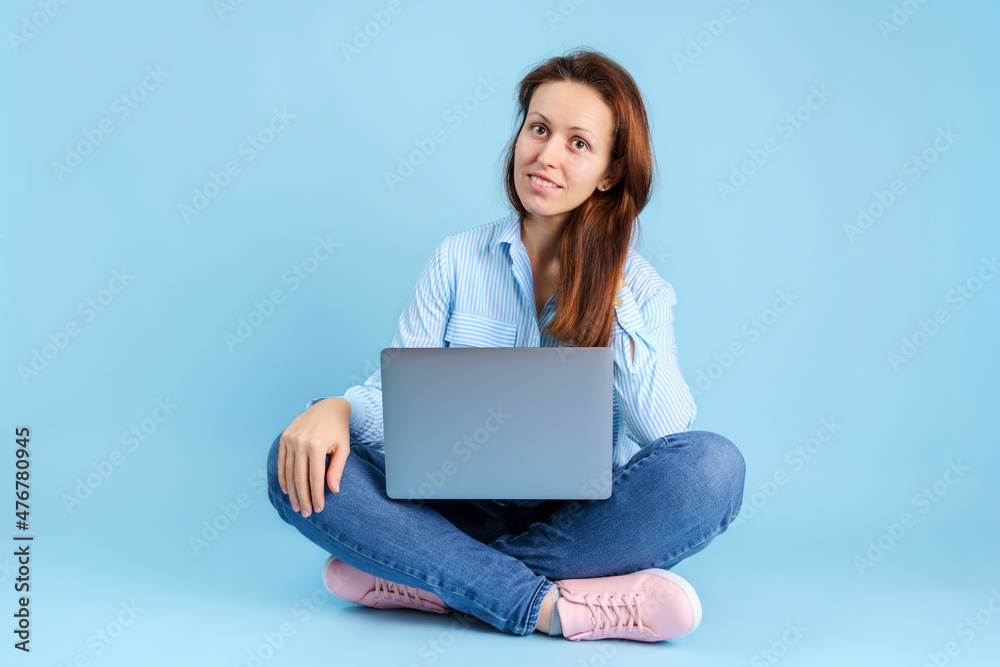 A beautiful freelancer girl with a laptop on her lap is sitting on a blue background A beautiful freelancer girl with a laptop on her lap is sitting on a blue background
