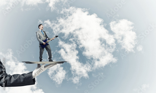 Businessman on metal tray playing electric guitar against blue sky background