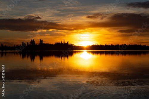 Orange sunset over lake with black silhouette tree line and clouds in the sky