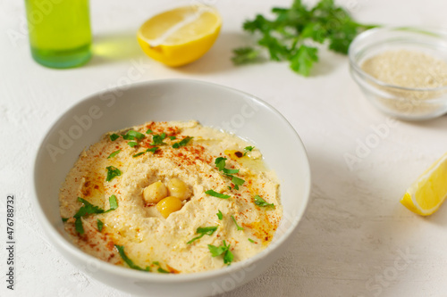 Hummus in a ceramic white bowl with paprika and parsley leaves and ingredients on a light background. Banner. A traditional Middle Eastern dish. Horizontal orientation. Vegetarian food concept.
