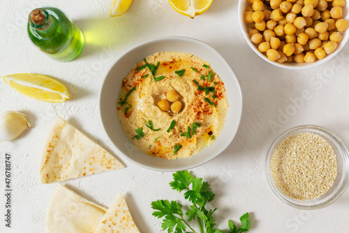 Hummus in a ceramic white bowl with paprika and parsley leaves and ingredients on a light background. A traditional Middle Eastern dish. Horizontal orientation. Vegan food concept. Top view