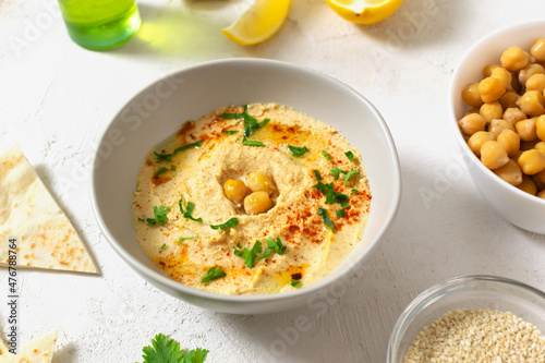 Hummus in a ceramic white bowl with paprika and parsley leaves and ingredients on a light background. A traditional Middle Eastern dish. Horizontal orientation. Healthy nutrition concept.