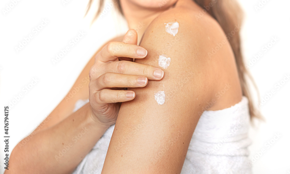 Fototapeta premium Closeup of young woman wrapped in towel applying body cream or lotion on shoulder after shower