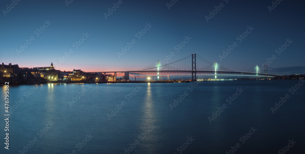 Two bridges against the sunset sky and city embankment in lights. Forth Road Bridge and Queensferry Crossing. South Queensferry, Scotland, United Kingdom