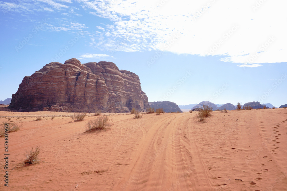 An empty road and a red weathered sandstone relief mountain in the Wadi Rum desert, rare dry bushes grow on the sand, Jordan An empty road and a red weathered sandstone relief mountain in the Wadi Rum desert, rare dry bushes grow on the sand, Jordan