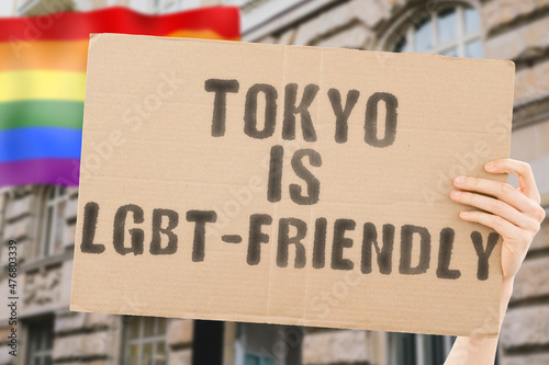 The phrase " Tokyo is LGBT-Friendly " on a banner in men's hand with blurred LGBT flag on the background. Human relationships. different. Diverse. liberty. Sexuality. Social issues. Society © AndriiKoval