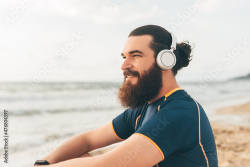 Young bearded fitness athletic man listening music resting looking aside at sunrise over the sea