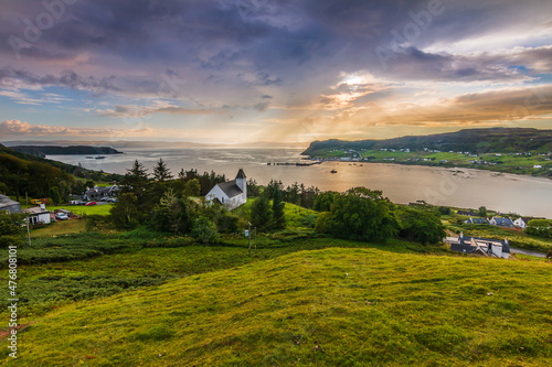 a place in Scotland on the Isle of Skye. Church on a hill with green meadow and trees. View over the landscape with sun rays on the horizon. Dramatic clouds in the sky at sunset