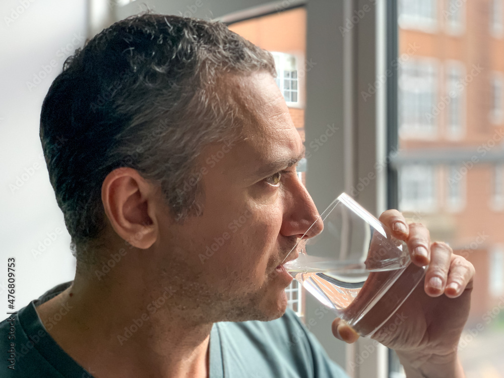 Man is drinking water from glass, staying by the window in Apartment. Middle aged man is drinking water to stay hydrated Thirsty man having hangover Pure life still water - obrazy, fototapety, plakaty 