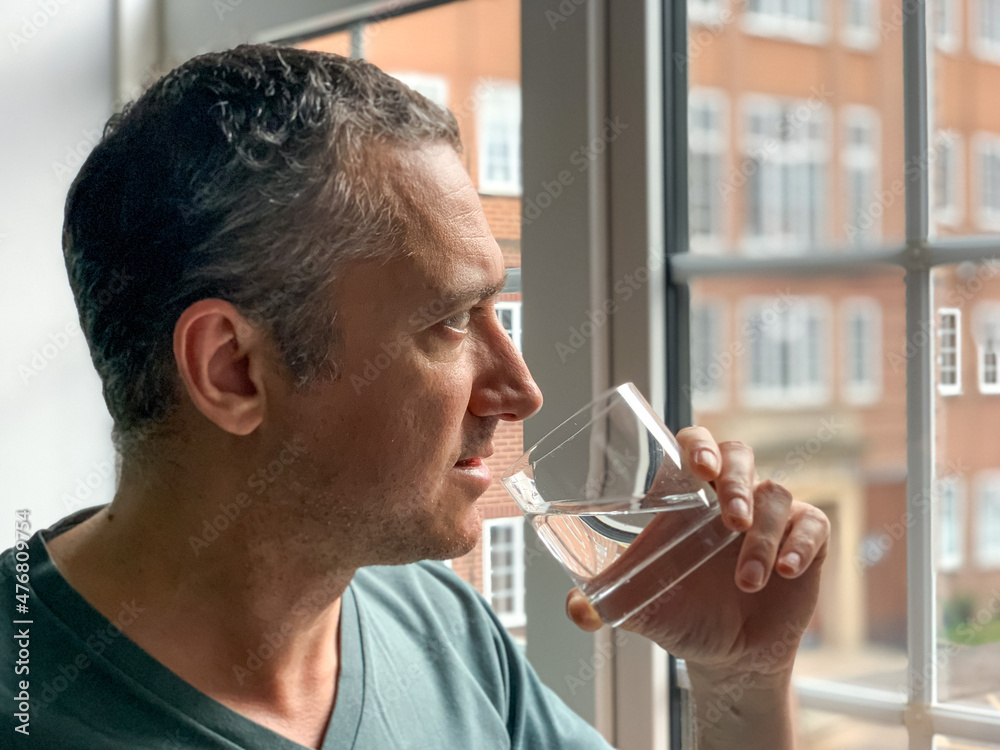 Man is drinking water from glass, staying by the window in Apartment. Middle aged man is drinking water to stay hydrated Thirsty man having hangover Pure life still water - obrazy, fototapety, plakaty 