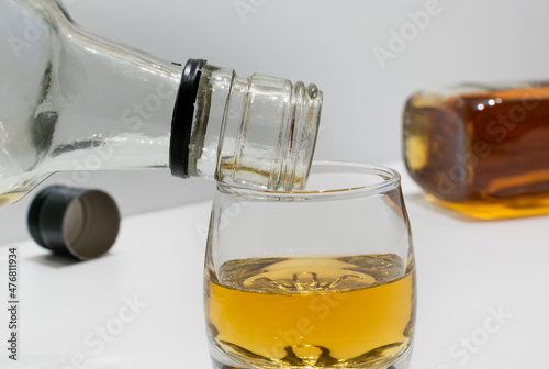Empty bottle on a glass with whiskey on white table close-up on blurred background of full bottle and cap