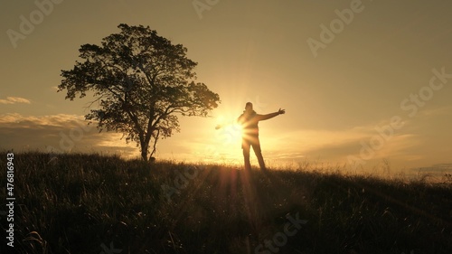 The silhouette of a traveler on a hill, a tourist rejoices, spins and raises his hands .. A backpacker walks up a hill past a lone tree in the sun. Outdoor adventure concept, sport lifestyle