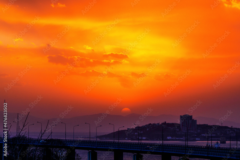 Bright sunset in Vladivostok. The red sun sets on the hills against the background of the road and the sea.