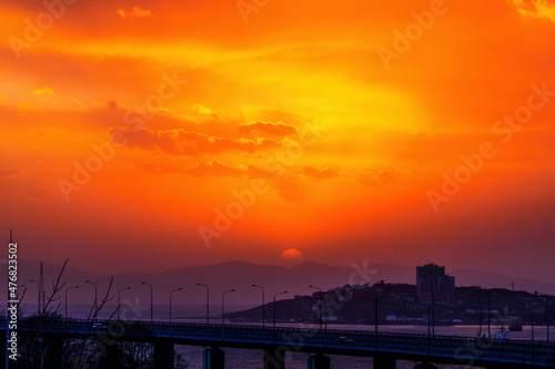 Bright sunset in Vladivostok. The red sun sets on the hills against the background of the road and the sea. © alexhitrov