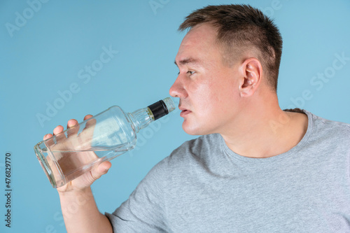 Portrait of a drunk man drinking strong alcohol from the throat of a bottle, close-up on a blue background