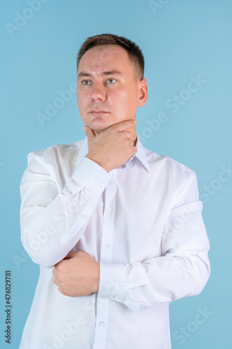 A man in a white shirt holds his hand to his chin and looks away standing on a blue background © Сергей Храмов