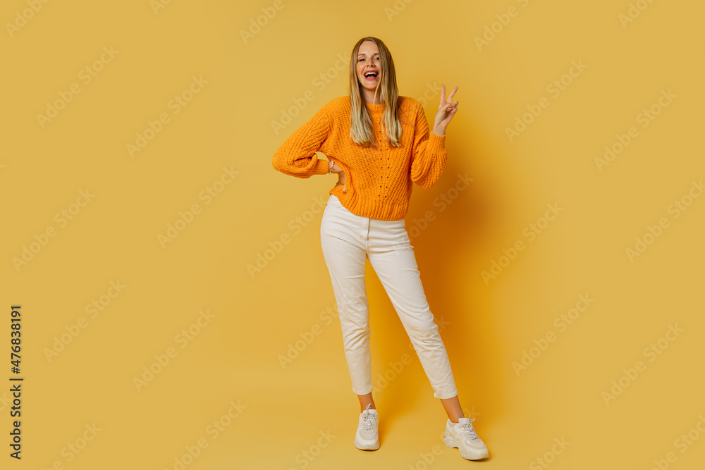 Obraz premium Blond woman with suprice face in orange stylish autumn sweater posing over yellow background in studio. Full lenght.